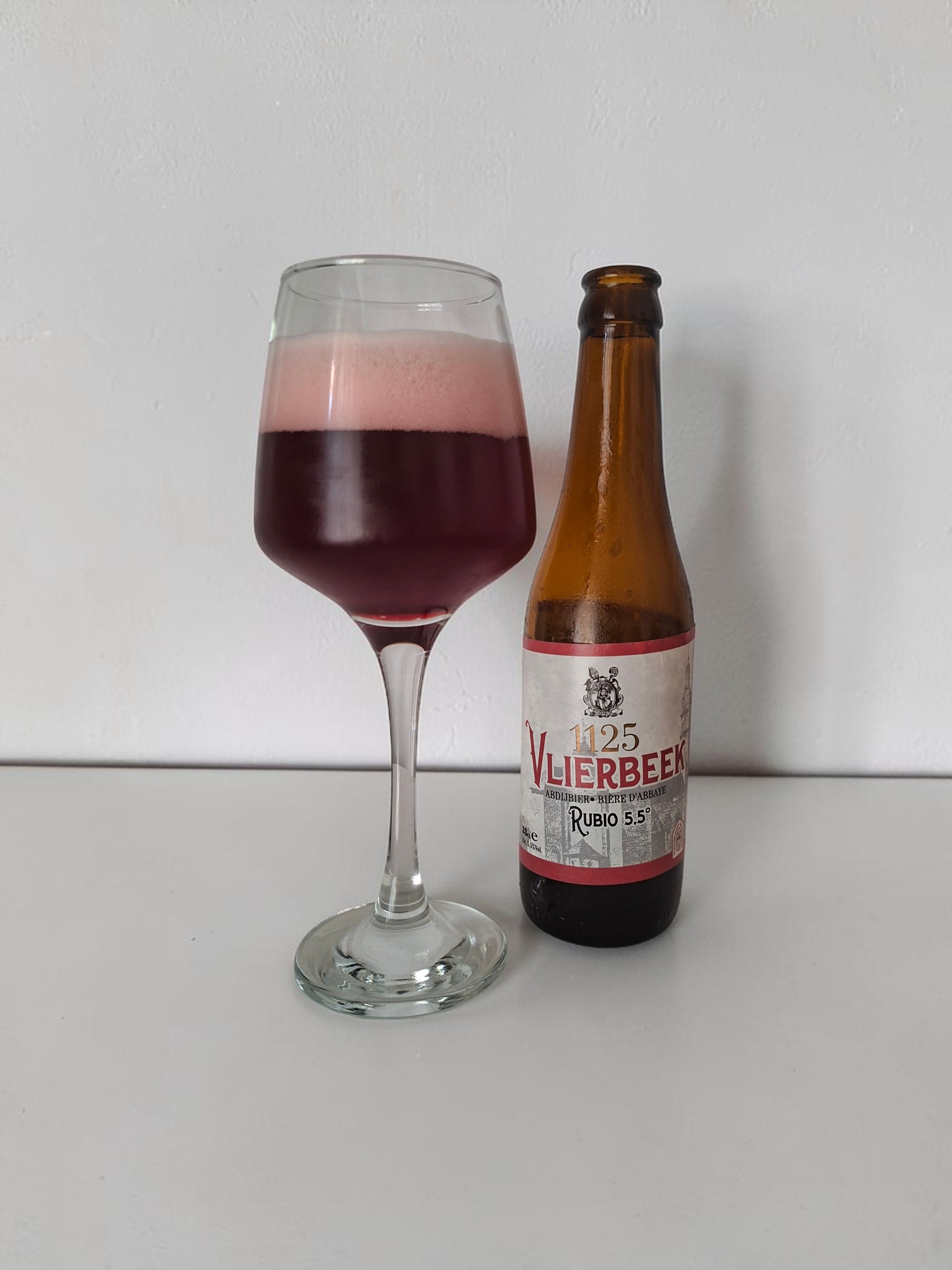Rubio poured into a glass beside its bottle, showing its deep red-amber color and pale pink foam head, on a clean white background